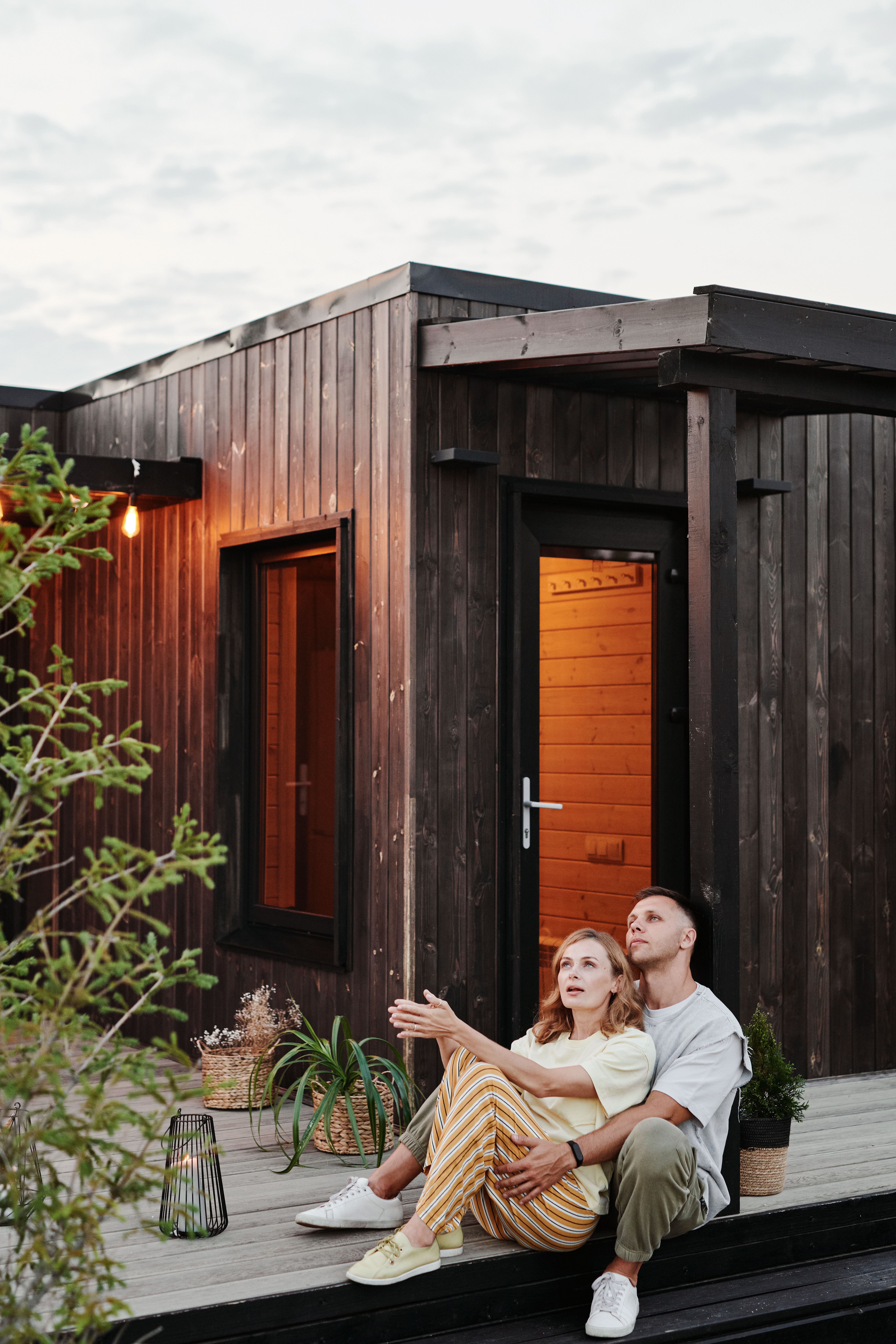 A couple sits together on a deck outside a wooden cabin, enjoying a peaceful evening.